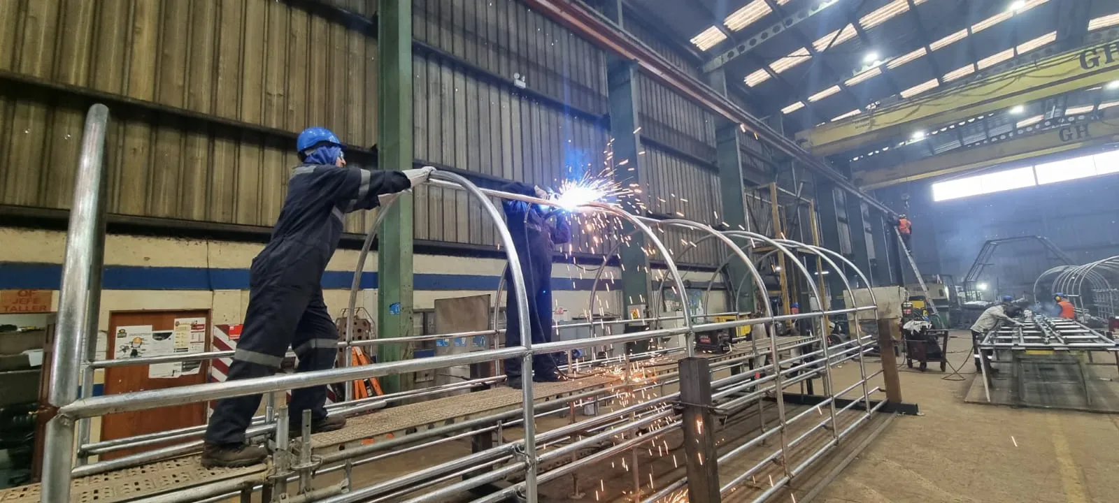 Dos operarios sueldan los arcos galvanizados de una cúpula sobre una plataforma dentro de la nave del taller, entre chispas